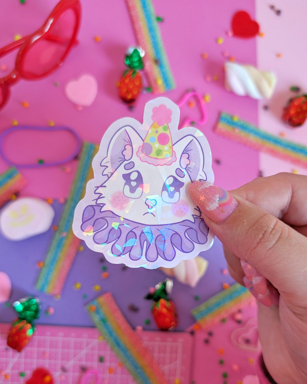 A hand with glittery pink nails holds a purple sticker featuring a stylized cat in a party hat and ruff. The backdrop is pink and purple with colourful candies.
