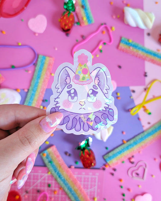 A hand with glittery pink nails holds a purple sticker featuring a stylized cat in a party hat and ruff. The backdrop is pink and purple with colourful candies.