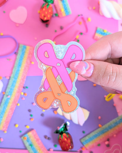 A hand with glittery pink nails holds a sticker of a 2 pairs of scissors, one pink and one orage, interlocking. The background is a colourful flatlay of pink and purple sheets decorated with candies.
