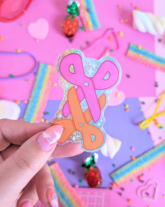 A hand with glittery pink nails holds a sticker of a 2 pairs of scissors, one pink and one orage, interlocking. The background is a colourful flatlay of pink and purple sheets decorated with candies.