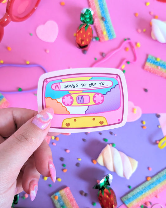 A hand with pink glittery nails holds a pastel cassette tape sticker with the text 'A SONGS TO CRY TO.' The backdrop is pink and purple with assorted candies.