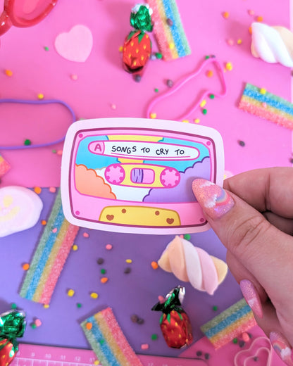 A hand with pink glittery nails holds a pastel cassette tape sticker with the text 'A SONGS TO CRY TO.' The backdrop is pink and purple with assorted candies.