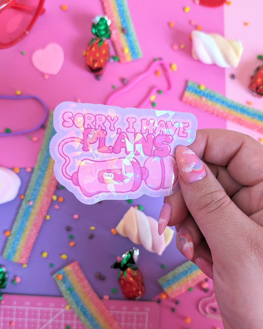 A hand with pink nails holds a sparkly sticker of a vibrator saying, "Sorry, I have plans," set over a pink and purple background with rainbow candies.