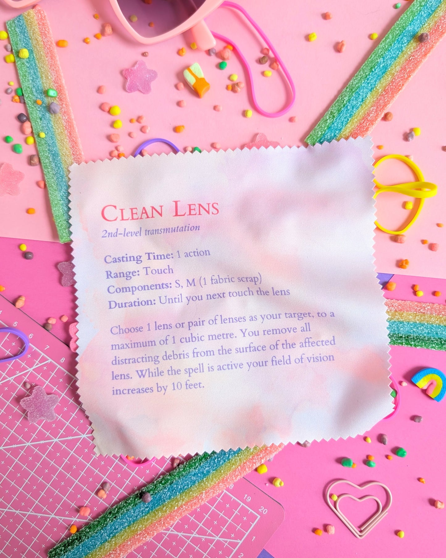 Lens wipe cloth with a Clean Lens spell on a pink background filled with colourful candies