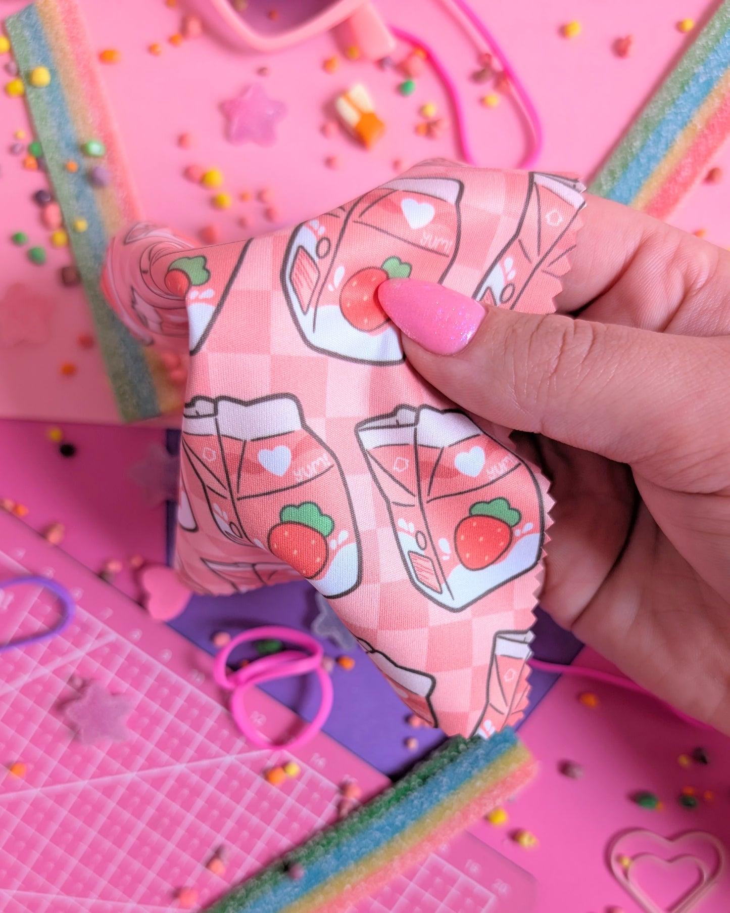 Hand with pink nails holding a draped lens wipe cloth with a strawberry milk pattern set against a pink and purple background filled with rainbow candies