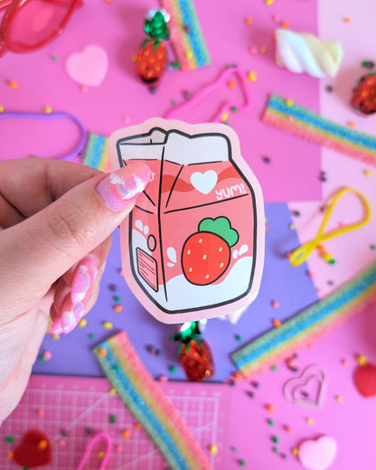 A hand with pastel pink nails holds a strawberry milk carton sticker against a colorful pink and purple background with candies scattered about.
