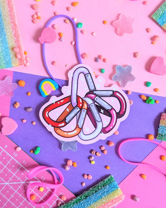 Sticker of carabiners in the colour of the lesbian pride flag overlapping in a semi-circle on a pink and purple background. Surrounded by rainbow candy.