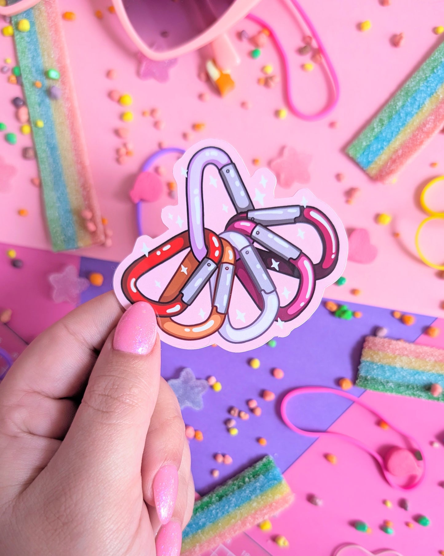 Hand with pink nails holding sticker of carabiners in the colour of the lesbian pride flag overlapping in a semi-circle on a pink and purple background. Surrounded by rainbow candy.