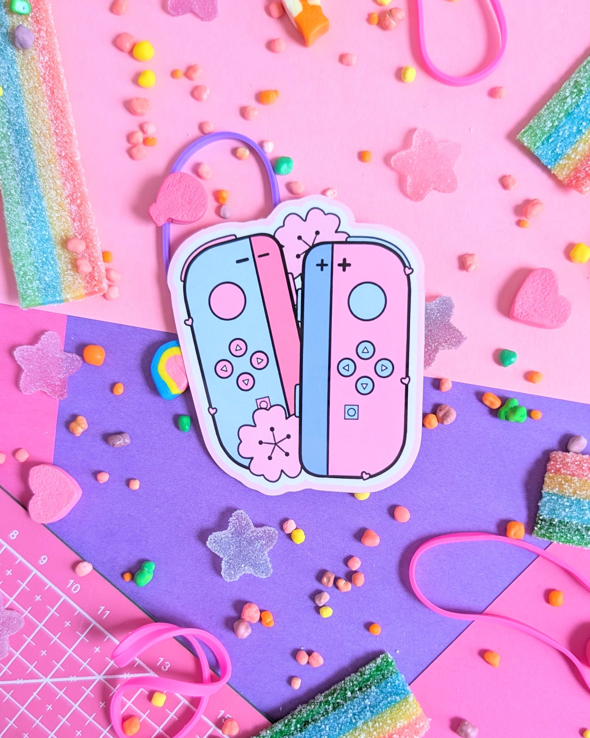 Sticker of pastel pink and blue gaming controllers on a vibrant pink and purple background. Surrounded by colourful candy