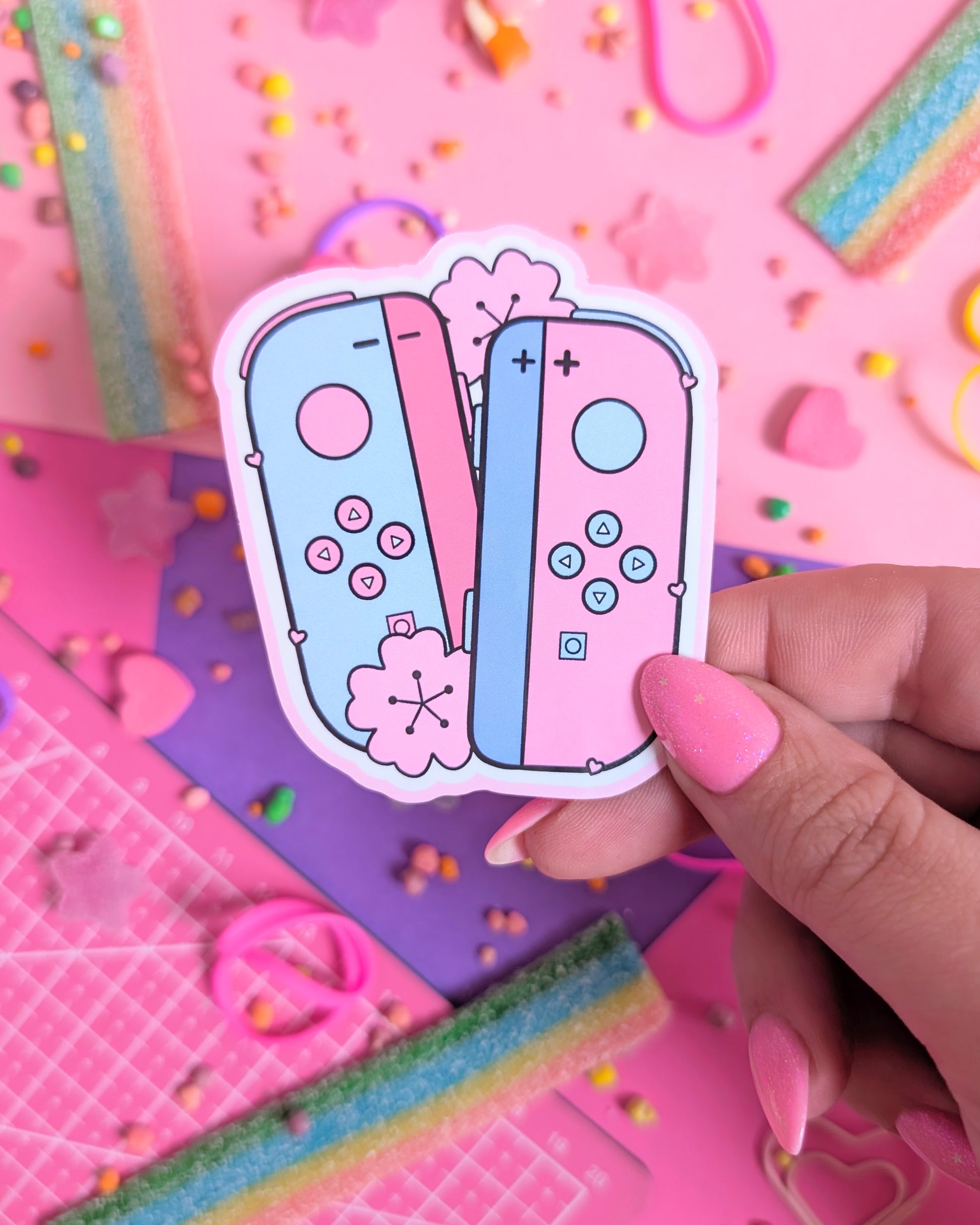 Hand with pink nails holding a sticker of pastel pink and blue gaming controllers on a vibrant pink and purple background. Surrounded by colourful candy