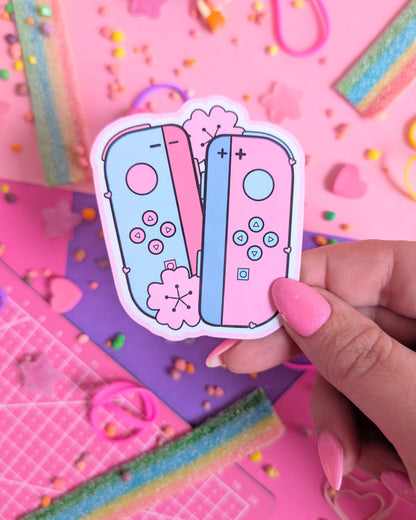 Hand with pink nails holding a sticker of pastel pink and blue gaming controllers on a vibrant pink and purple background. Surrounded by colourful candy
