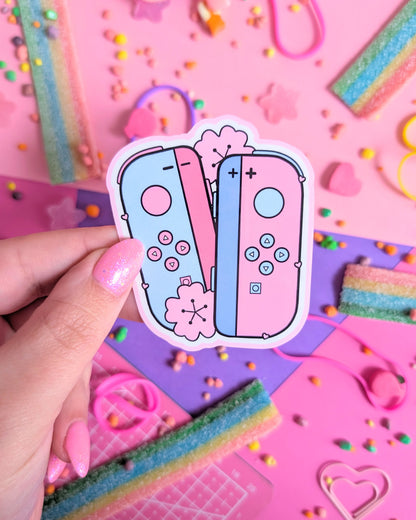 Hand with pink nails holding a sticker of pastel pink and blue gaming controllers on a vibrant pink and purple background. Surrounded by colourful candy