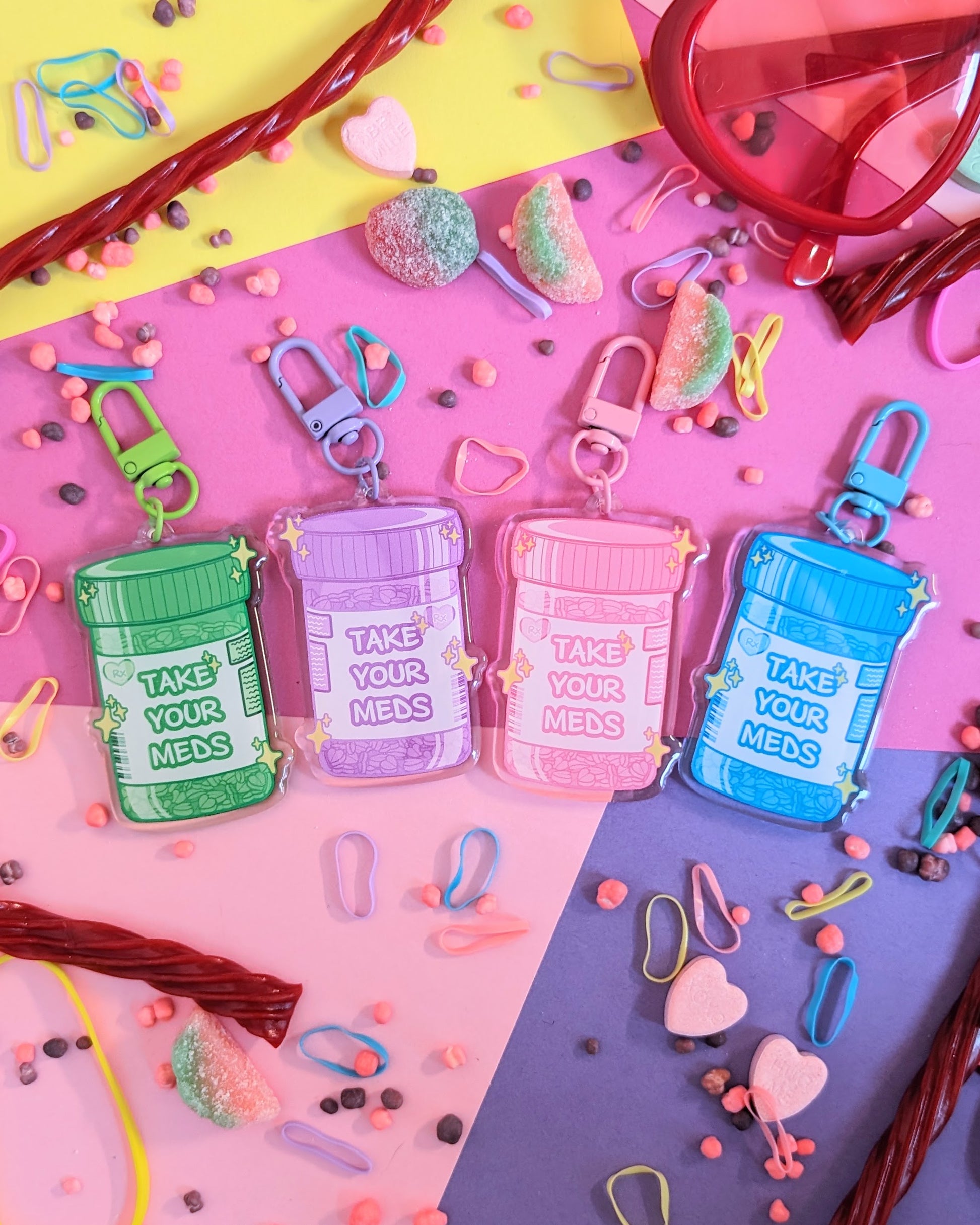 Four colourful keychains shaped like prescription bottles with "Take Your Meds" text against a pink and purple background with rainbow candies