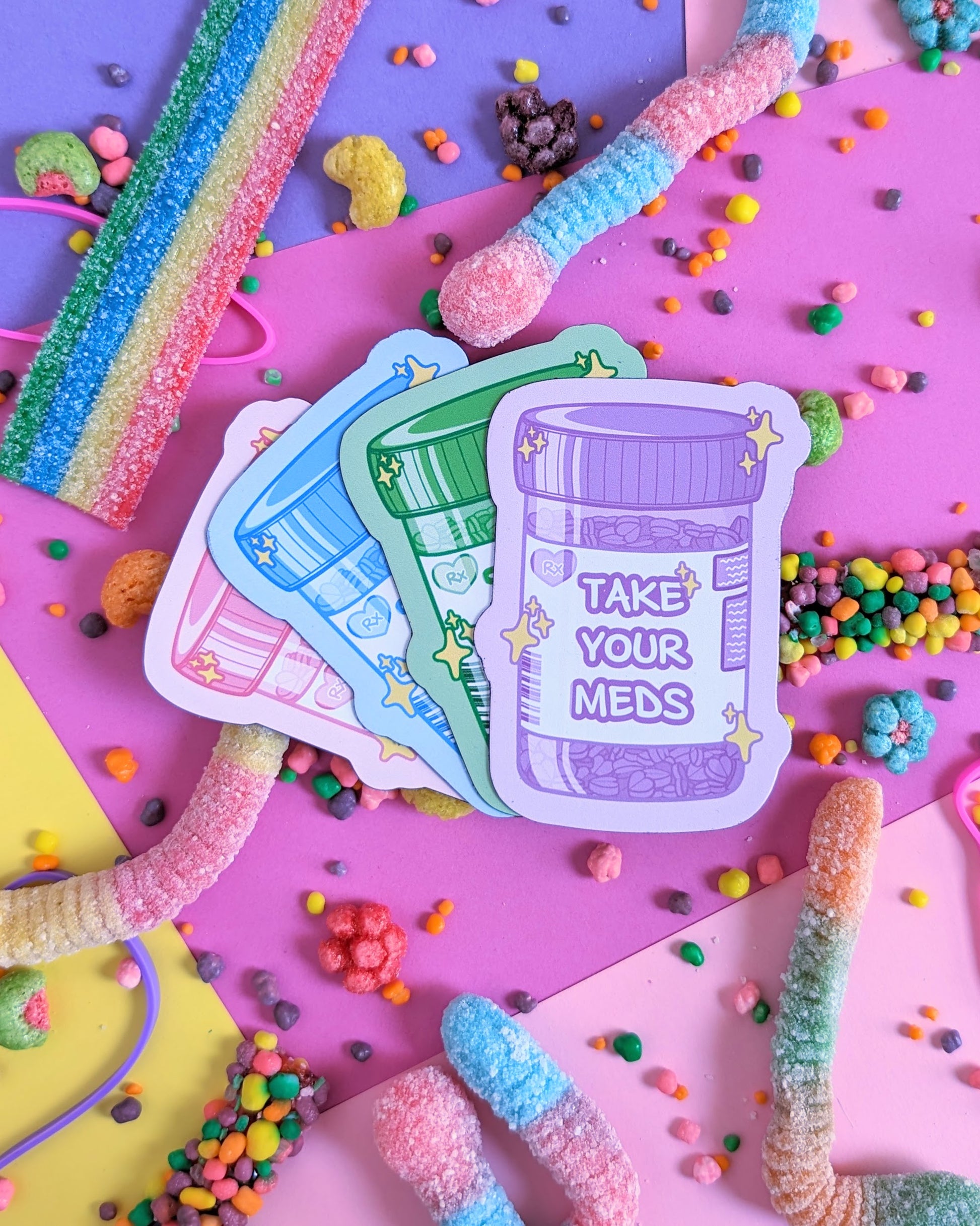 Four magnets shaped like prescription bottles that say "Take Your Meds" set against a pink and purple background with rainbow candies