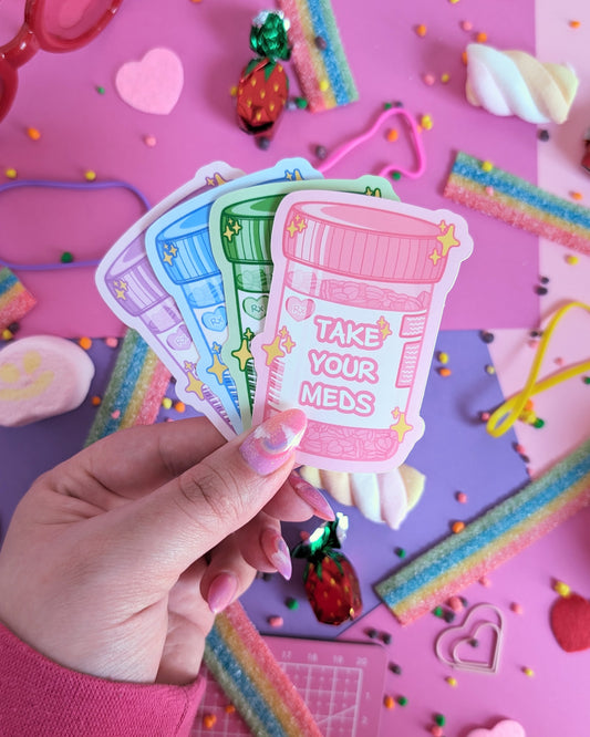 A hand with pink nails holds four stickers shaped like prescription bottles saying "Take Your Meds", set against a pink and purple background with rainbow candies