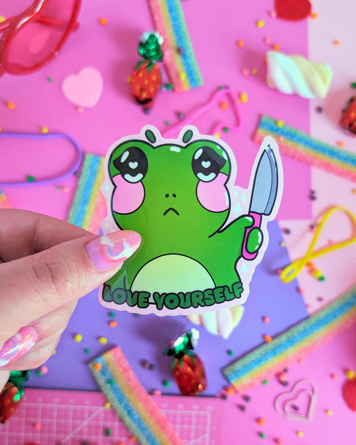 A hand with glittery pink nails holds a green sticker featuring a stylized frog holding a knife with the caption 'LOVE YOURSELF.' The backdrop is pink and purple with colourful candies.