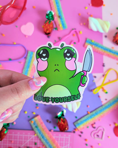 A hand with glittery pink nails holds a green sticker featuring a stylized frog holding a knife with the caption 'LOVE YOURSELF.' The backdrop is pink and purple with colourful candies.