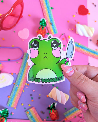 A hand with glittery pink nails holds a green sticker featuring a stylized frog holding a knife with the caption 'LOVE YOURSELF.' The backdrop is pink and purple with colourful candies.