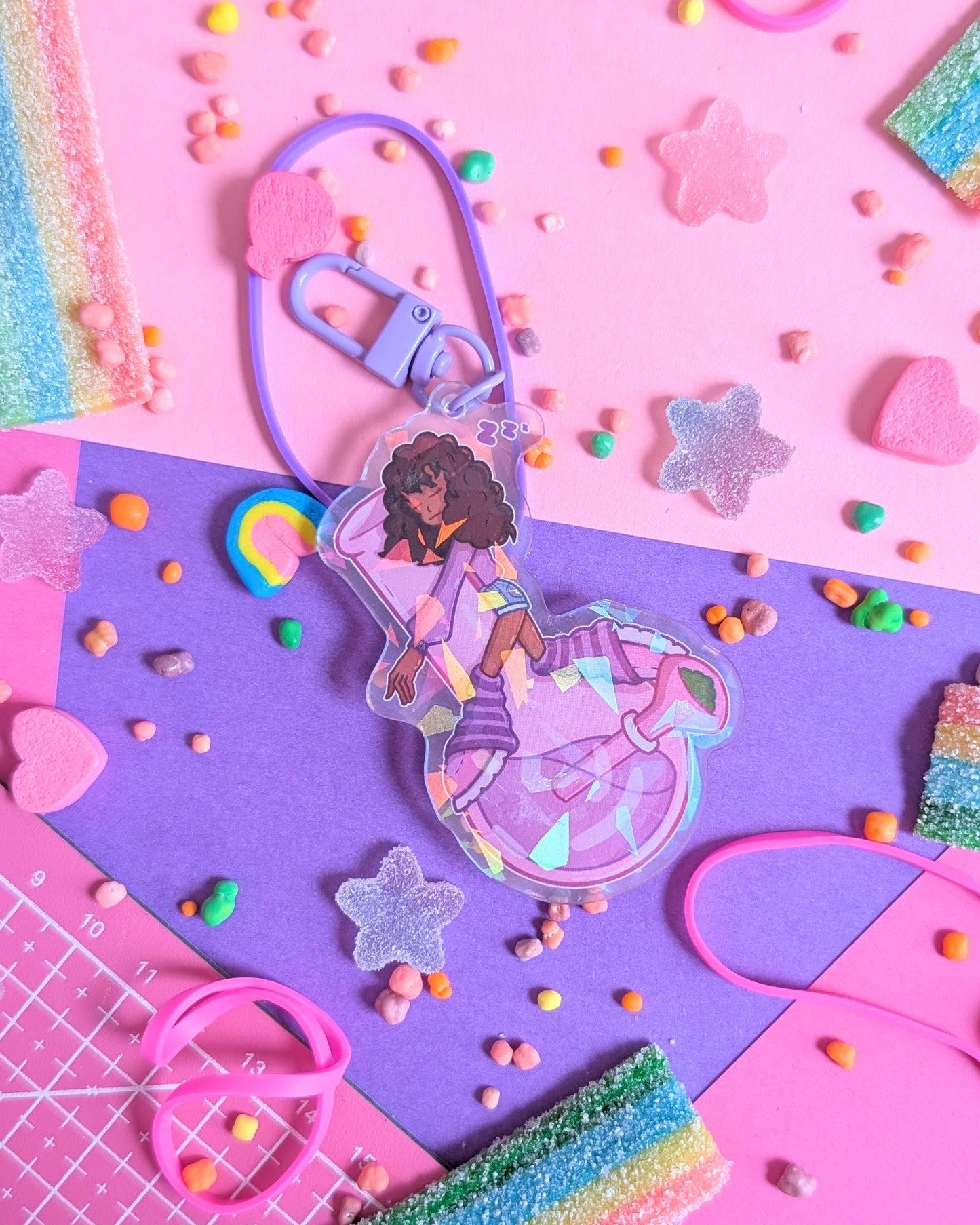 Keychain charm of a Black girl with curly hair sleeping on a purple bong set against a pink and purple background with rainbow candies.