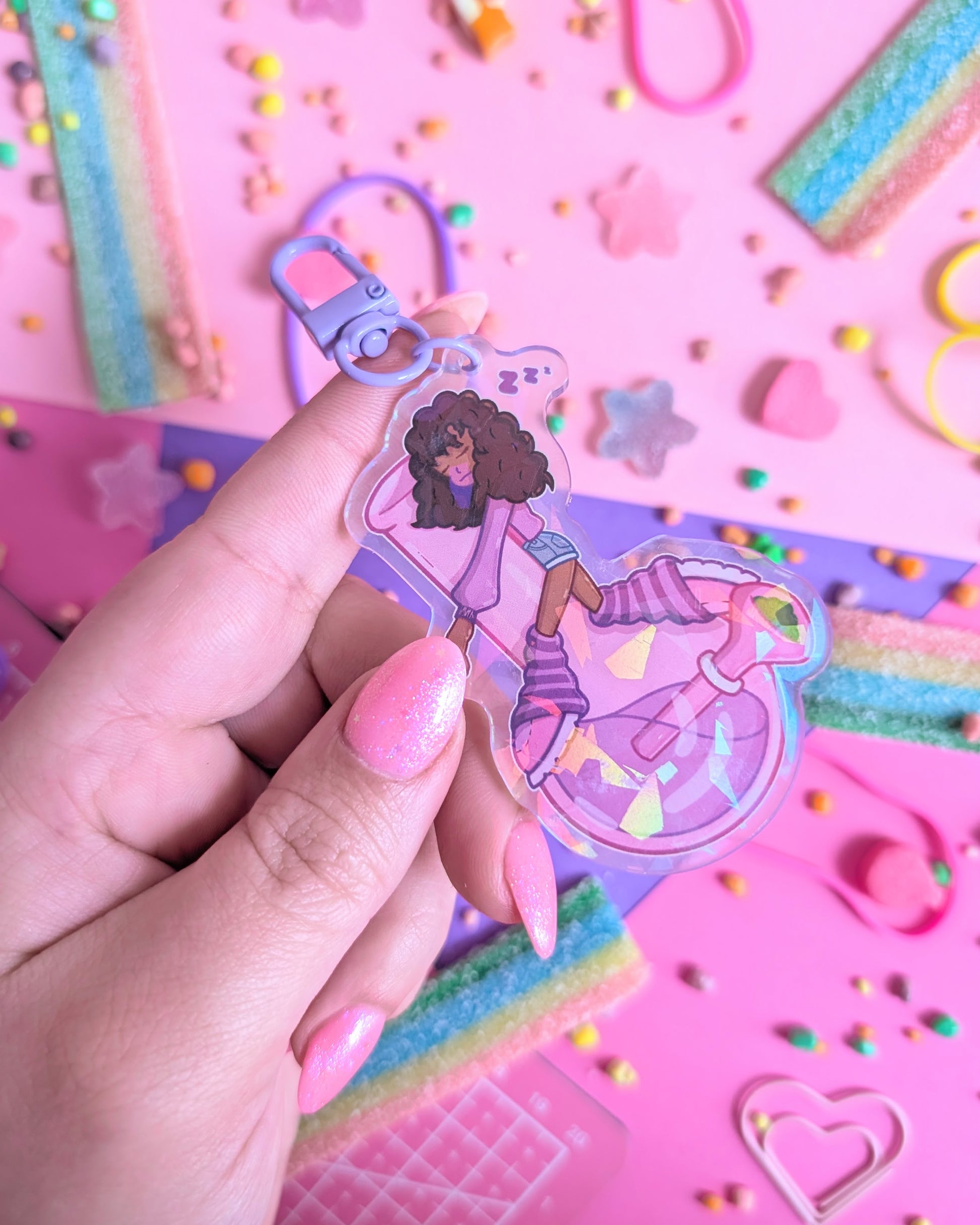 Hand with pink nails holding a keychain charm of a Black girl with curly hair sleeping on a purple bong set against a pink and purple background with rainbow candies.