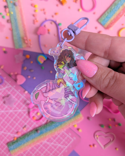 Hand with pink nails holding a keychain charm of a Black girl with curly hair sleeping on a purple bong set against a pink and purple background with rainbow candies.