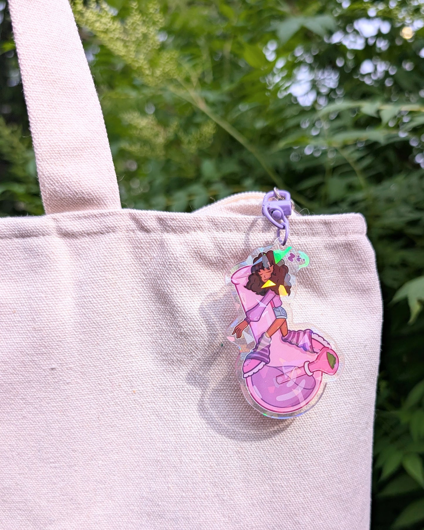Keychain charm of a Black girl with curly hair sleeping on a purple bong hanging from a blank canvas tote set against a background of green leaves