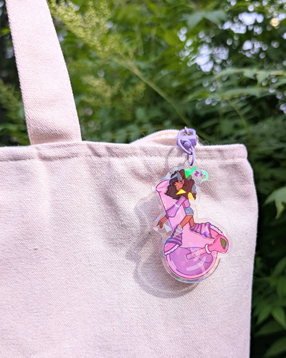 Keychain charm of a Black girl with curly hair sleeping on a purple bong hanging from a blank canvas tote set against a background of green leaves