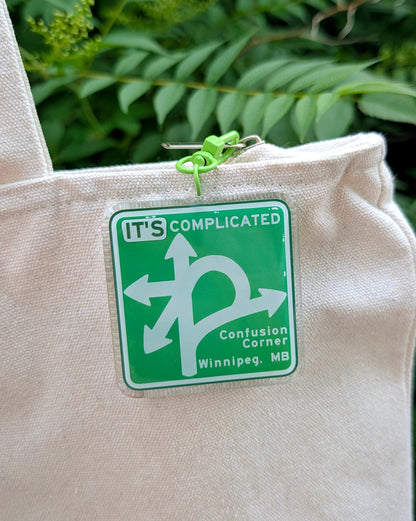 Beige tote bag with a green and white keychain charm of a road sign sign reading 'It's Complicated Confusion Corner Winnipeg, MB' attached.